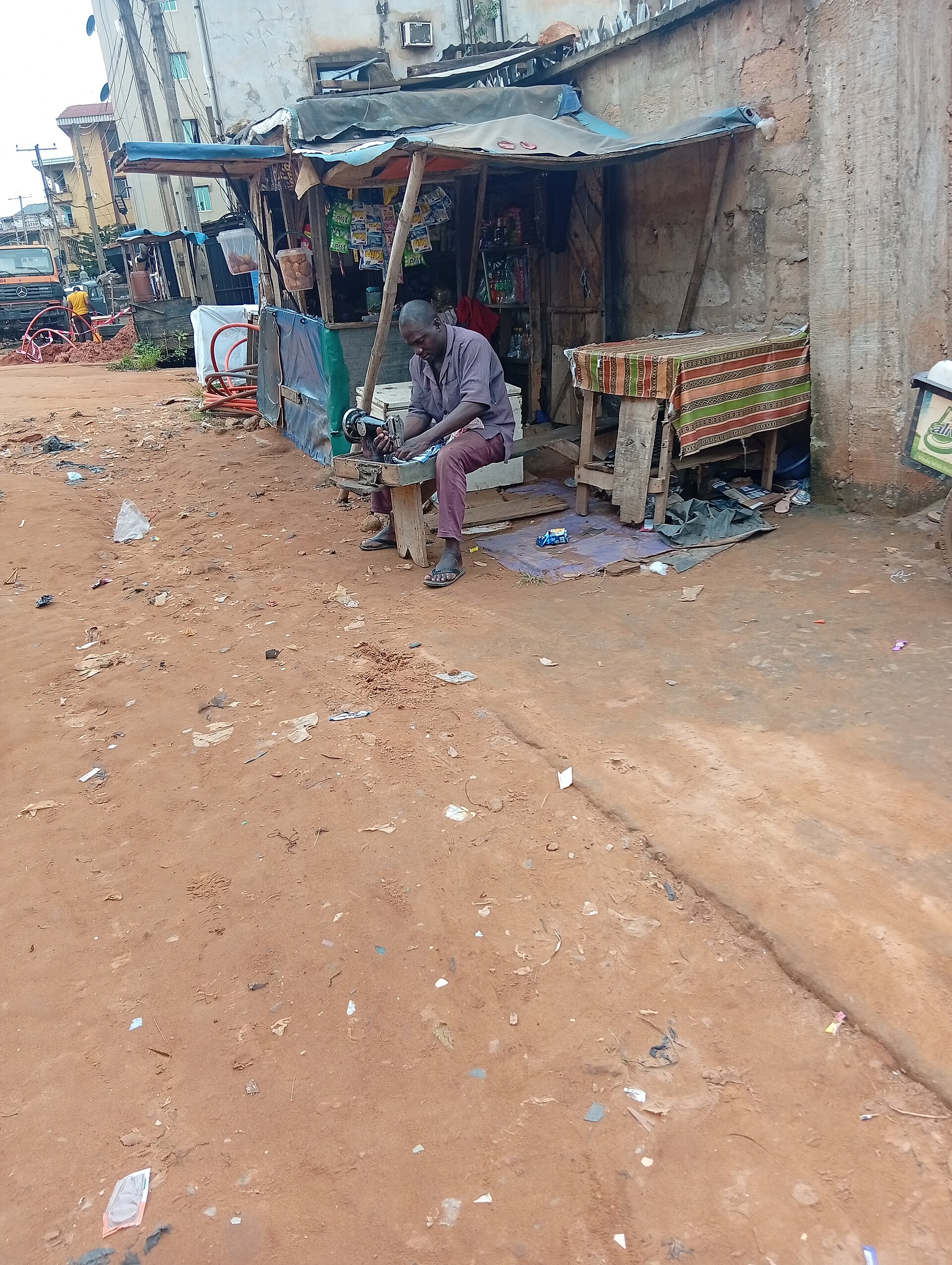 A man sewing cloth on the street with a sewing machine, providing tailoring service in an outdoor workspace. The scene captures everyday cra