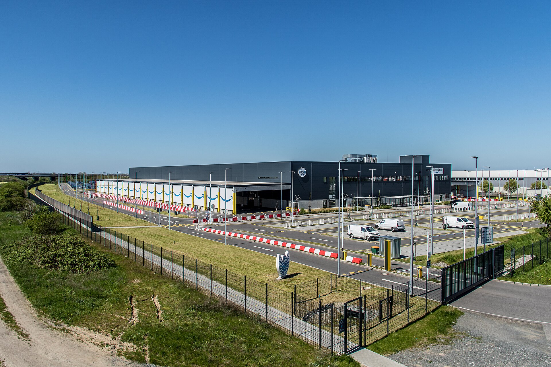 Wide view of the Amazon logistics centre DSY1 in Germany: a long warehouse, car park and fence under a clear sky