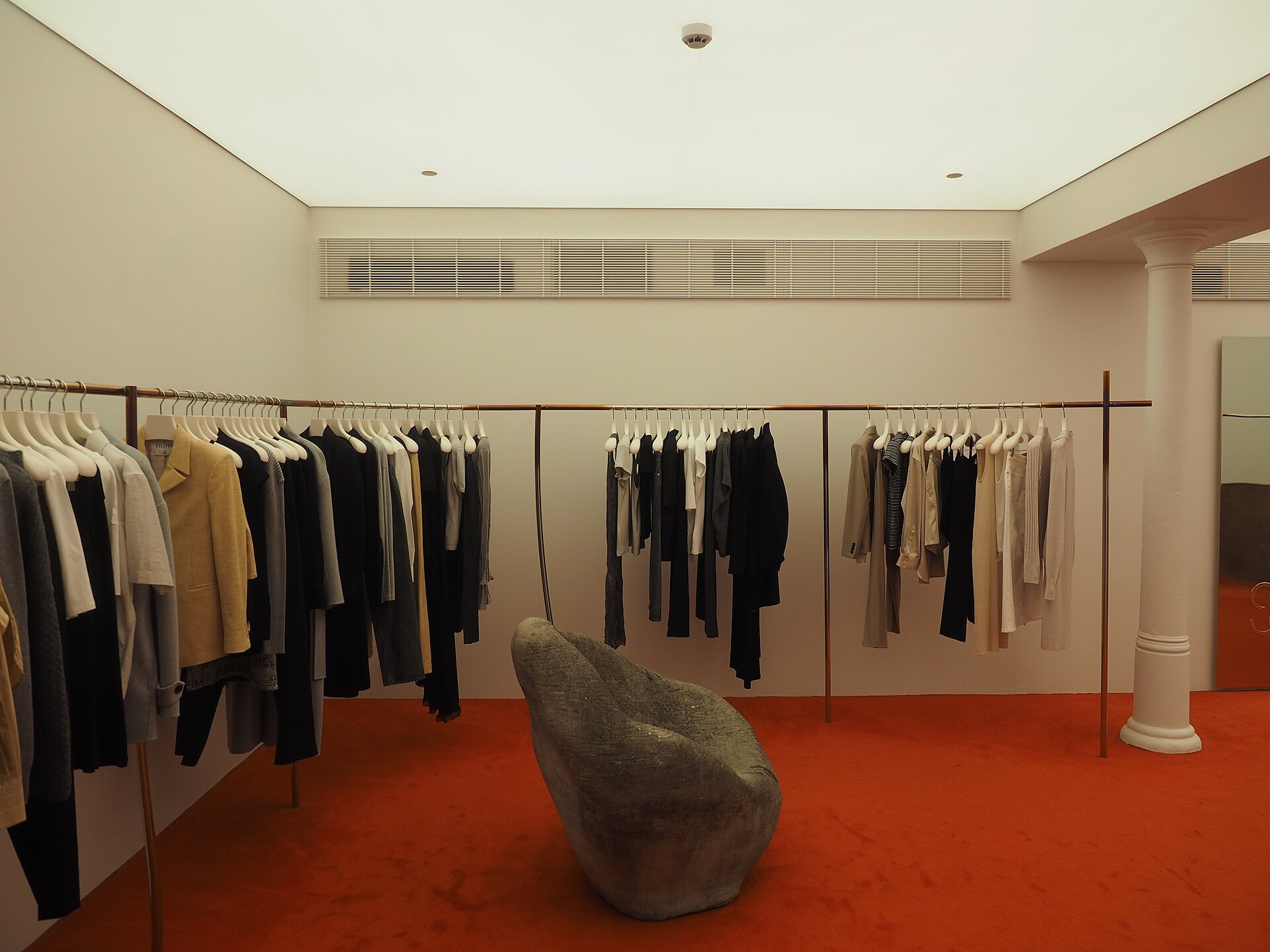 Clothes including jackets, trousers and dresses hanging on a rack with a grey armchair in front of them at the 25th anniversary of the Kämp