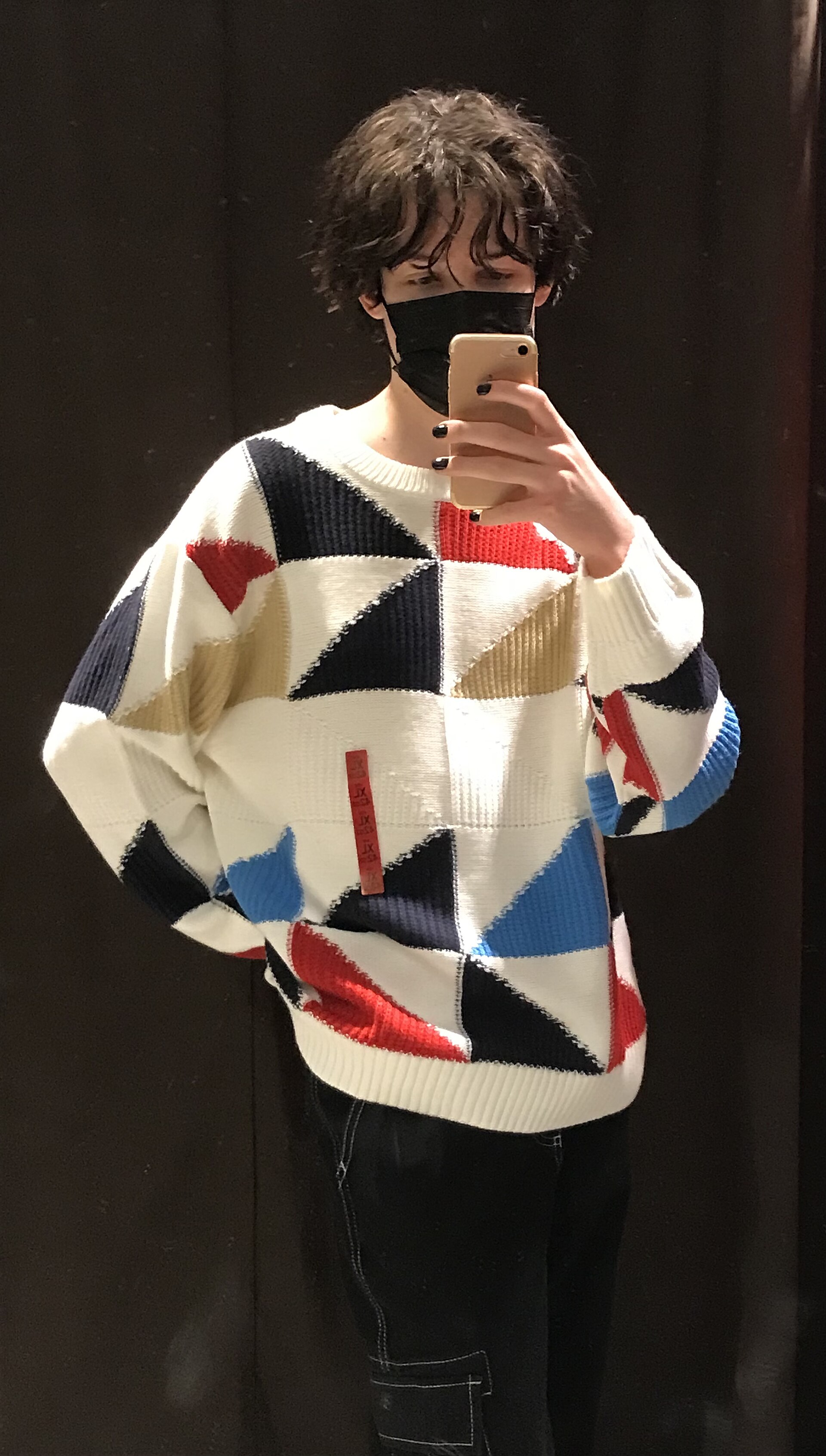 A person wearing a knitted sweater with blue and red triangles, and black pants that are either jeans or cargos (don’t really know). There i